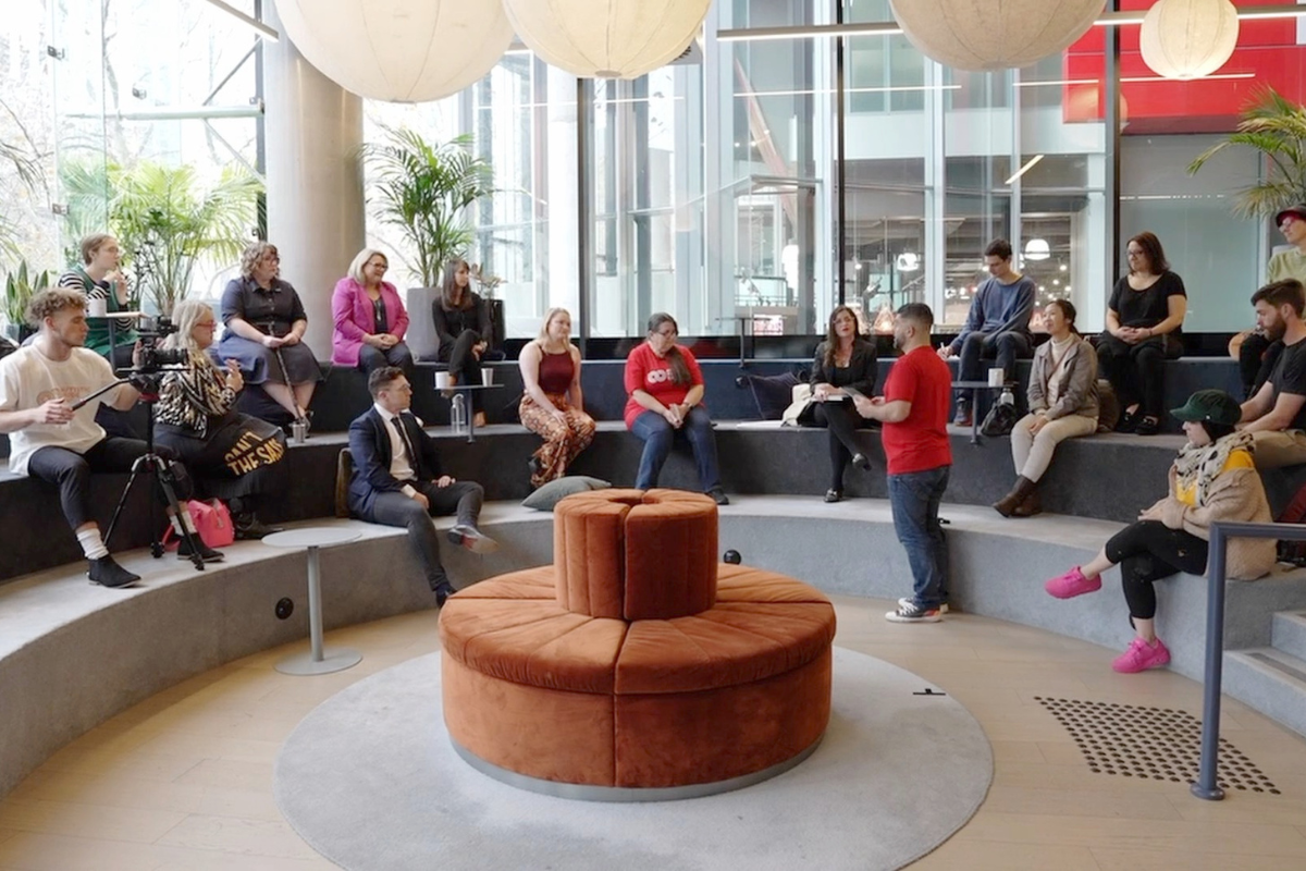 A group of people gathered in a modern, open space, attentively listening to a speaker in a red shirt, with a videographer recording the session.